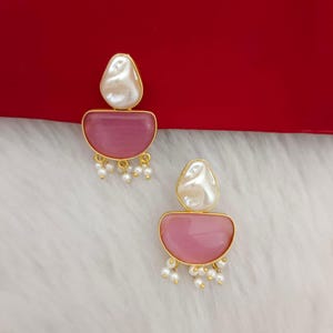 Könnte beinhalten: Ein Paar goldene Ohrringe mit rosa und weißen Steinen. Die Ohrringe haben einen tropfenförmigen weißen Stein auf einem halbkreisförmigen rosa Stein. Die Ohrringe sind mit kleinen weißen Perlen verziert.