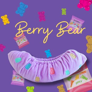 May include: A lavender-colored plush item with embroidered gummy bear designs, surrounded by colorful gummy bear candies and candy packets on a purple background. The text "Berry Bear" is written in a cursive font.