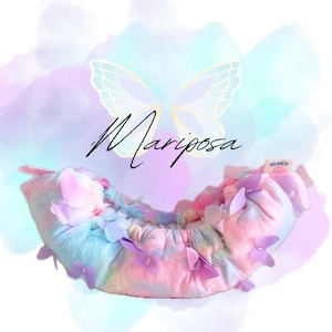 May include: A pastel-colored hair accessory with butterfly accents. The product is decorated with lavender and pink butterflies. The word "Mariposa" is written in cursive. The background is a watercolor wash of pastel colors.