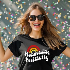 Puede incluir: Una mujer con una camiseta negra con un gráfico de arcoíris y el texto "Normalizar la positividad" en blanco. Está sonriendo y señalando con ambas manos.