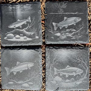 Könnte beinhalten: Set aus vier quadratischen Schieferuntersetzern, jeweils mit einem detaillierten Bild eines Fisches unter Wasser geätzt. Die Fische sind von Wasserpflanzen, Felsen und einem Baumstamm umgeben. Die Untersetzer sind dunkelgrau.