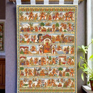 Op de afbeelding: Een kleurrijke, religieuze rol met een lijst, die verschillende goden en sc&egrave;nes afbeeldt. Het kunstwerk heeft ingewikkelde details, levendige kleuren en traditionele Indiase motieven. De rol is gemonteerd op een houten staaf met witte stoffen banden om op te hangen. Het thema is spiritueel en cultureel.