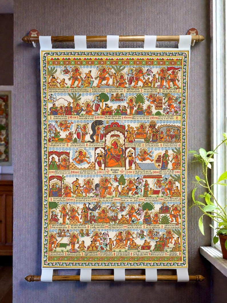 Op de afbeelding: Een kleurrijke, religieuze rol met een lijst, die verschillende goden en sc&egrave;nes afbeeldt. Het kunstwerk heeft ingewikkelde details, levendige kleuren en traditionele Indiase motieven. De rol is gemonteerd op een houten staaf met witte stoffen banden om op te hangen. Het thema is spiritueel en cultureel.