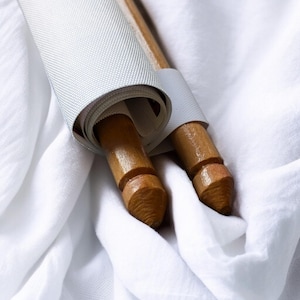 Op de afbeelding: Twee houten scrollstokken met een opgerolde witte stof. De stokken hebben een lichtbruine afwerking en worden bij elkaar gehouden met een witte band. De stof is lichtgrijs van kleur en is opgerold rond de stokken. De achtergrond is een witte stof.