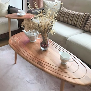 May include: Two oval wooden coffee tables with a light wood finish. The larger table has a unique design with concentric circles around the edge. A vase with dried flowers sits on the larger table, along with a teapot and a creamer.