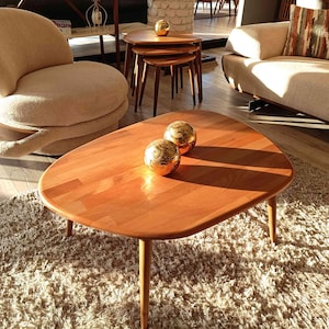 A wooden coffee table with a rounded oval top and four legs. The table is sitting on a fluffy white rug. Two gold decorative spheres are placed on the table.