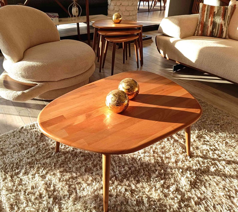 A wooden coffee table with a rounded oval top and four legs. The table is sitting on a fluffy white rug. Two gold decorative spheres are placed on the table.