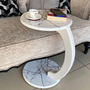 May include: White marble side table with a curved base and a round top. The table has a white marble top with black veining and a white base.
