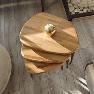 May include: Three wooden nesting tables with a natural wood finish. The top table has a round, gold-colored decorative object on it.