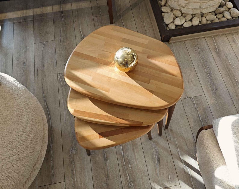 May include: Three wooden nesting tables with a natural wood finish. The top table has a round, gold-colored decorative object on it.