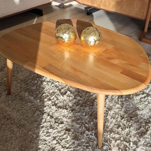 May include: A light wood oval coffee table with two gold decorative spheres on top. The table has three legs and is sitting on a light brown shag rug.