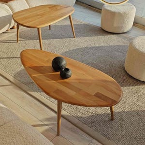 May include: Two wooden oval coffee tables with a natural wood finish. The larger table has a decorative, oval shape and sits on four legs. The smaller table has a more traditional rectangular shape and sits on four legs. Both tables are on a beige rug with two white poufs.