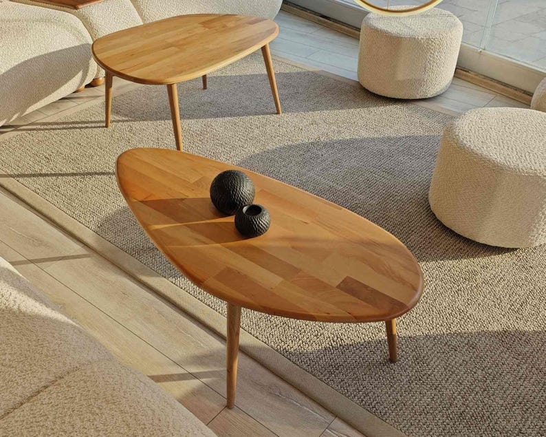May include: Two wooden oval coffee tables with a natural wood finish. The larger table has a decorative, oval shape and sits on four legs. The smaller table has a more traditional rectangular shape and sits on four legs. Both tables are on a beige rug with two white poufs.