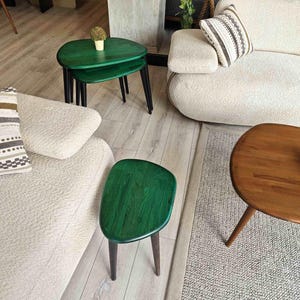 Three green wooden side tables with rounded edges and black metal legs. The tables are arranged in a living room setting with a white couch and a brown wooden coffee table.
