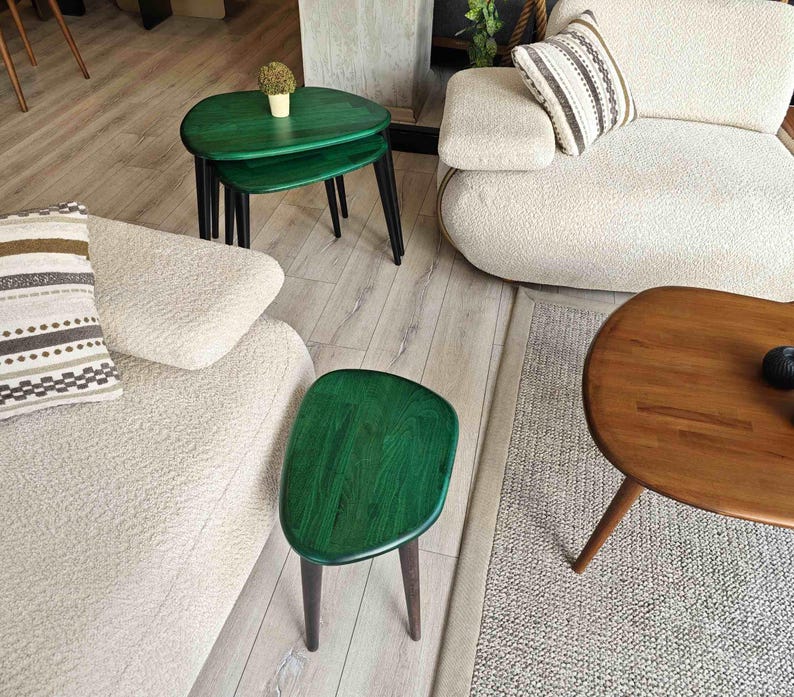 Three green wooden side tables with rounded edges and black metal legs. The tables are arranged in a living room setting with a white couch and a brown wooden coffee table.