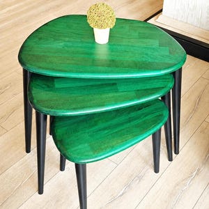 May include: A set of three nesting tables with a vibrant green tabletop and black legs. The tables are different sizes, with a small plant in a white pot on the largest table. The tables have a modern, minimalist design.