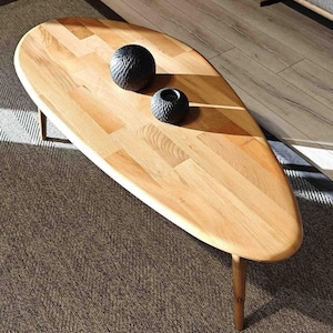 May include: A light wood oval coffee table with a natural wood finish. The table has a unique shape and is supported by three thin legs. Two black decorative objects are placed on the table.