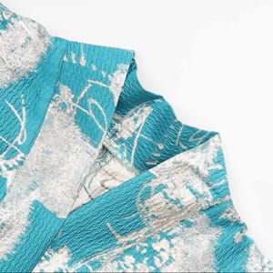 May include: Close-up of a teal blue fabric with a silver metallic pattern. The fabric has a textured surface.
