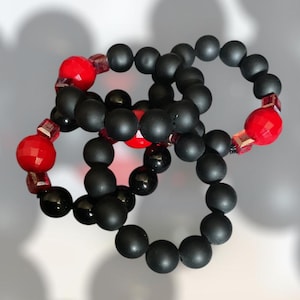 Puede incluir: Tres pulseras de cuentas negras con cuentas rojas de acento. Las pulseras están hechas de cuentas negras mate y presentan cuentas rojas facetadas y cuentas de acento rojas y transparentes más pequeñas. Las pulseras están entrelazadas.