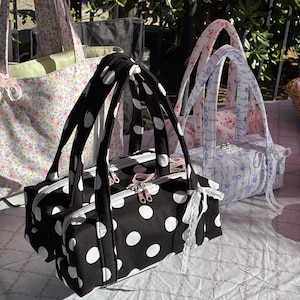 Peut inclure: Trois sacs en tissu avec anses. Le sac au premier plan est noir à pois blancs avec un ruban en dentelle blanche. Derrière, un sac blanc présente un motif de rubans bleus, et un grand cabas à fleurs est visible en arrière-plan.