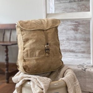 May include: A vintage brown canvas backpack sits on top of a white ceramic crock with a blue number "5" on the side. The crock is covered with a beige linen fabric. The backpack has a flap with a buckle closure.