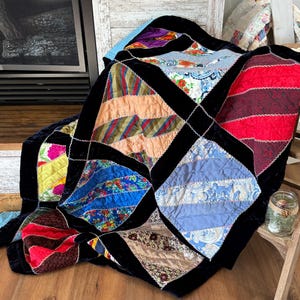 May include: A patchwork quilt made from various fabrics in shades of blue, red, green, and yellow. The quilt is made of squares with black velvet trim. The quilt is folded and draped over a wooden chair.