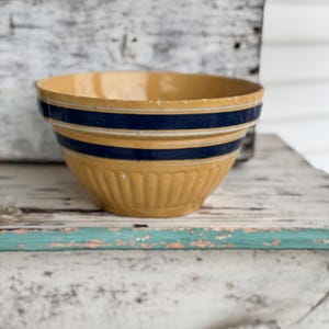 May include: A vintage yellow ceramic mixing bowl with two navy blue stripes. The bowl has a ribbed exterior and a slightly chipped rim, suggesting age and use. This farmhouse-style bowl is ideal for kitchen decor or baking.