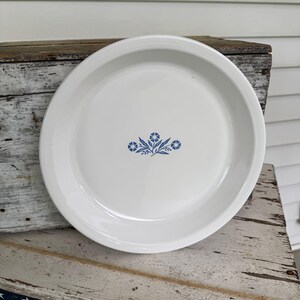 May include: White ceramic pie plate with a blue floral design in the center. The plate has a wide rim and is set on a rustic wooden surface. The floral design is a cluster of small blue flowers and leaves.