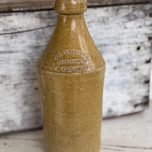 May include: A vintage, tan-colored stoneware bottle with a rounded body and a narrow neck. The bottle has embossed text that reads "C.S. TWITCHELL, PRESTON TO, & WHITMORE". The bottle is displayed on a white wooden surface.
