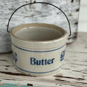 May include: A vintage butter crock with a wire handle. The off-white ceramic container features blue lettering that spells "Butter" and decorative accents. The crock is sitting on a distressed, painted wooden surface.