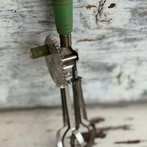 May include: Vintage hand-crank egg beater with a green wooden handle. The beater is made of metal and features a gear-driven mechanism and a circular blade. The handle is painted green. The beater is designed for mixing ingredients in the kitchen.