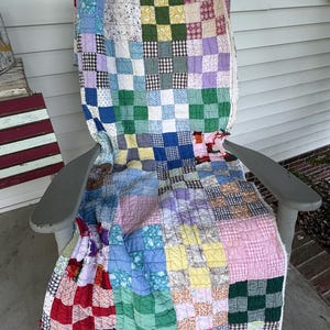 May include: A patchwork quilt featuring a grid of multi-colored fabric squares in various patterns including gingham, floral, and solid colors. The quilt is draped over a gray wooden rocking chair, displaying visible stitching throughout the fabric.
