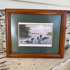 May include: A framed print featuring a lake scene with two adult loons and a chick swimming near reeds. The artwork is enclosed in a dark green mat and a polished medium-brown wooden frame, presenting a detailed illustration of the birds.