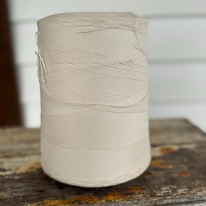 May include: A large spool of off-white thread, tightly wound and cylindrical in shape. The thread appears to be cotton or a similar natural fiber. The spool is resting on a weathered wooden surface, suggesting a craft or sewing supply.
