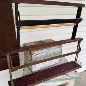 May include: An antique wooden and metal paper cutter with a dark brown finish. The paper cutter has three horizontal bars and a wooden dowel. The frame is supported by metal brackets. The paper cutter is sitting on a wooden box.