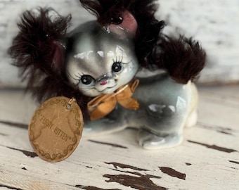 May include: A vintage ceramic kitten figurine with a gray and white body, dark fur accents, and a gold bow. The figurine has a tag that reads "Furry Kitten". The kitten is posed on a distressed white wooden surface.
