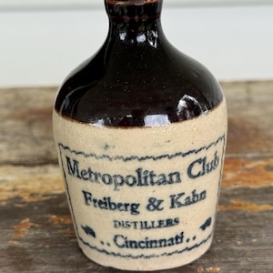 May include: A small, antique stoneware bottle with a dark brown top and a tan base. The bottle features the text "Metropolitan Club Freiberg & Kahn Distillers Cincinnati" in blue lettering. The bottle is likely a vintage collectible.