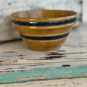 May include: A vintage stoneware bowl with a tan base and two dark blue stripes. The bowl has a slightly flared rim and a classic, rustic design. The bowl is likely used for serving or display.
