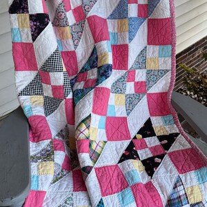 May include: A patchwork quilt featuring a repeating block pattern composed of various fabric squares in shades of pink, white, yellow, blue, and black. The quilt includes diverse prints such as plaid, floral, and gingham, finished with a pink patterned border.