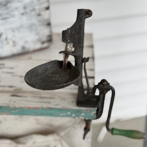 May include: An antique cast iron scale with a small, round tray and a green wooden handle. The scale is mounted on a wooden surface with peeling paint. The overall color scheme is muted with a rustic aesthetic.