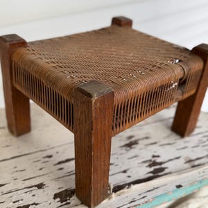 May include: A small, square wooden footstool with a woven rattan seat. The legs and frame are made of dark brown wood. The footstool is a vintage piece, showing signs of wear and age.