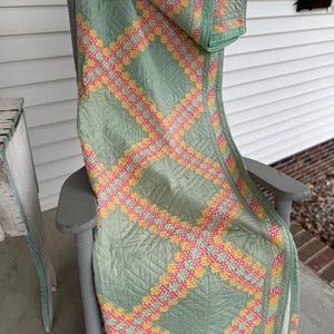 May include: A vintage quilt draped over a gray rocking chair. The quilt features a light green background with a repeating pattern of pink, yellow, and white polka dots. The quilt's edges are trimmed with a matching green border.