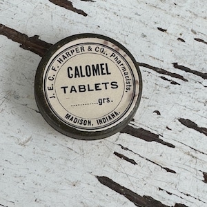 May include: A vintage, round metal tin with a white label featuring the text "CALOMEL TABLETS" and the name "J.E.C. F. HARPER & CO., Pharmacists." The tin is dark green with a distressed appearance, and the text "MADISON, INDIANA" is also visible.