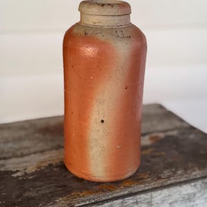 May include: A cylindrical, antique stoneware bottle with a light beige top and a reddish-orange body. The bottle has a narrow neck and a slightly textured surface. The number "1L" is visible on the side. The bottle is sitting on a weathered wooden surface.