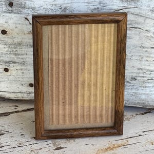 May include: A rectangular picture frame with a dark brown wood grain finish. The frame features a clear glass front covering a piece of light tan, vertically corrugated cardboard backing that fills the entire interior display area.
