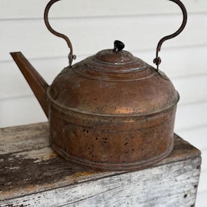 May include: A vintage copper kettle with a curved handle and a black wooden grip. The kettle has a spout and a lid with a small black knob. The kettle is sitting on a weathered wooden surface.