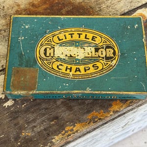 May include: A rectangular teal metal tin with a distressed finish. The lid features a central yellow oval emblem with the text Little Chancellor Chaps in black lettering. The side of the tin also displays the text Little Chancellor Chaps.