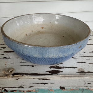 May include: A large, antique blue and white ceramic bowl. The bowl has a wide rim and a textured, patterned band around the middle. The interior is off-white with signs of age and use. A rustic, vintage kitchenware item.