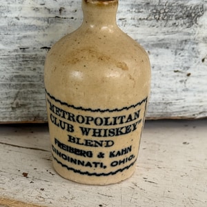 May include: A small, antique stoneware whiskey bottle with a tan glaze. The bottle has a handle and a label with the text "Metropolitan Club Whiskey Blend" and the name "Freiberg & Kahn, Cincinnati, Ohio" in blue lettering.