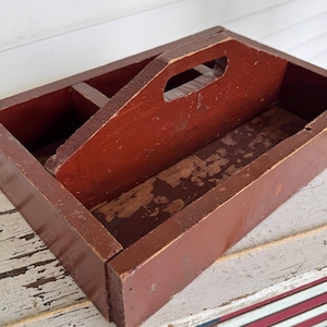 May include: A rectangular wooden caddy with a dark reddish-brown finish and a central handle. The caddy features a divided interior compartment with visible wear and paint chipping on the bottom surface, showcasing the natural wood grain underneath.
